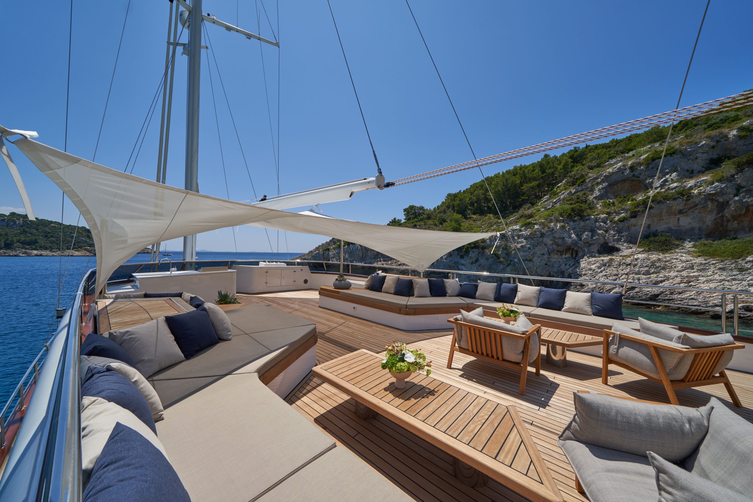 MARALLURE Luxury yacht deck with cushioned seating, wooden tables, and sunshade sails. The yacht is anchored near a rocky coastline with green trees under a clear blue sky.