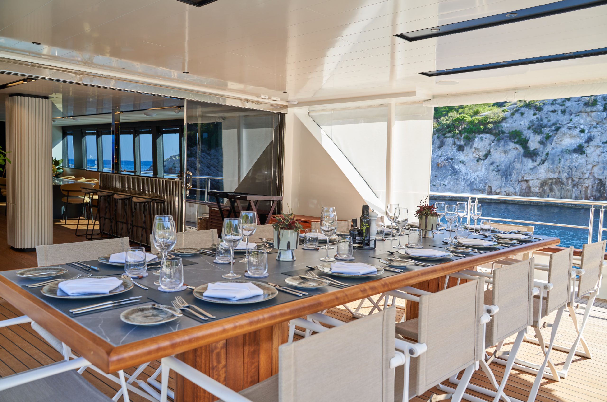 MARALLURE Elegant outdoor dining area on a yacht, set with plates, glasses, and cutlery for a meal. The table overlooks the sea and rocky coastline, with sunlight streaming in and modern seating around the wooden table.