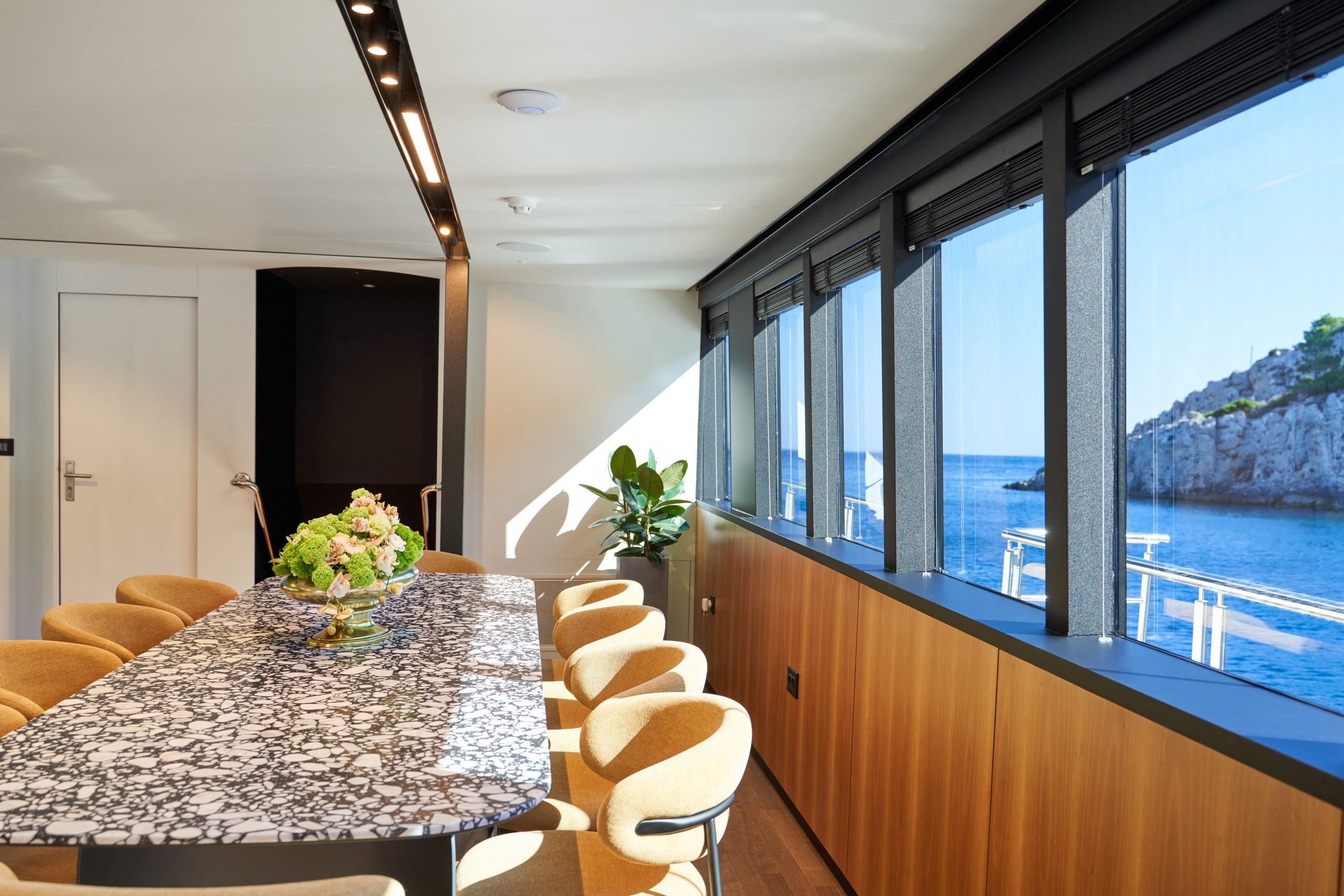 MARALLURE A modern dining room with a long marble table, tan chairs, a floral centerpiece, wood-paneled walls, large windows with ocean views, and natural sunlight streaming in.