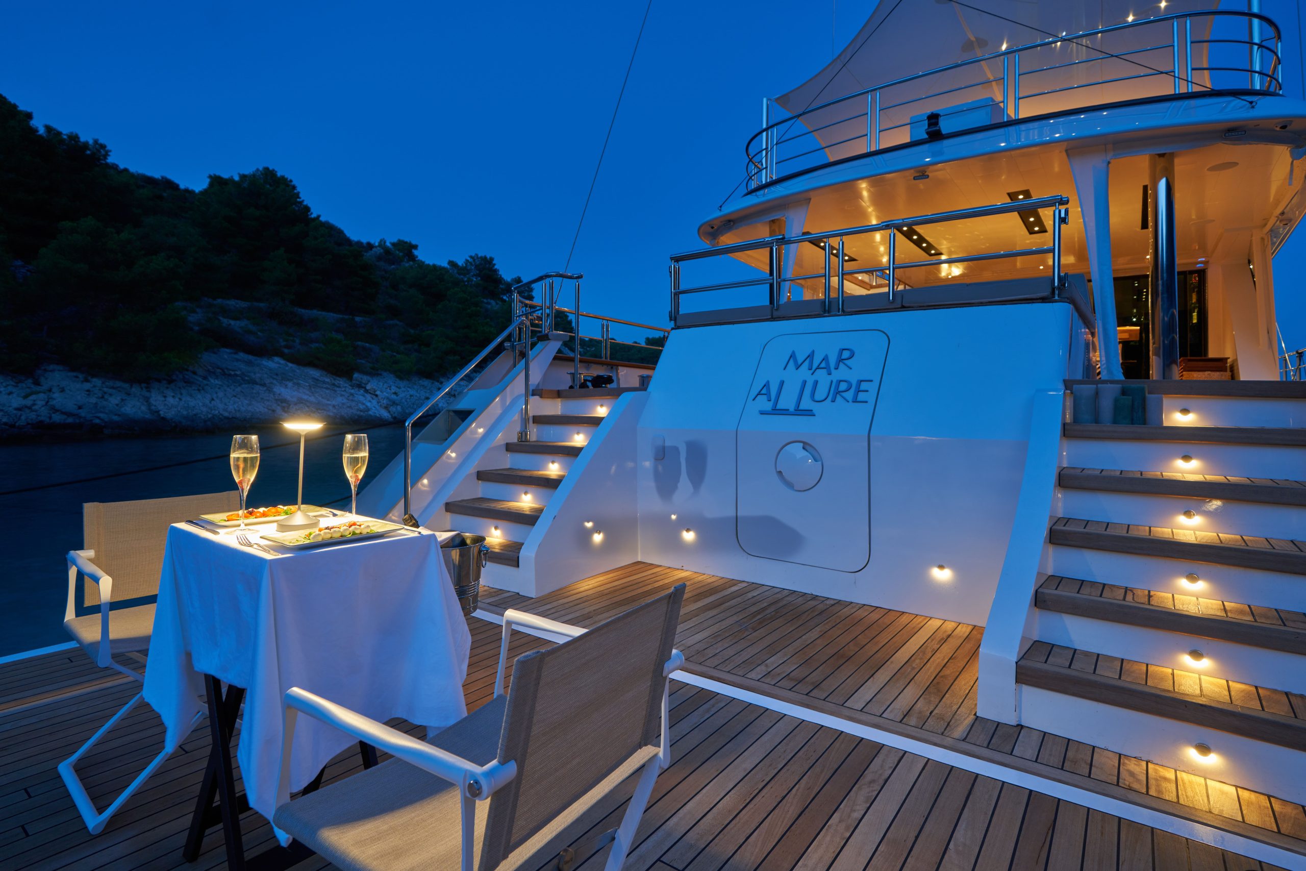 MARALLURE A romantic dinner table with two chairs, champagne glasses, and a lit lamp is set on the deck of a luxury yacht at dusk, with warm lights and water and trees in the background.