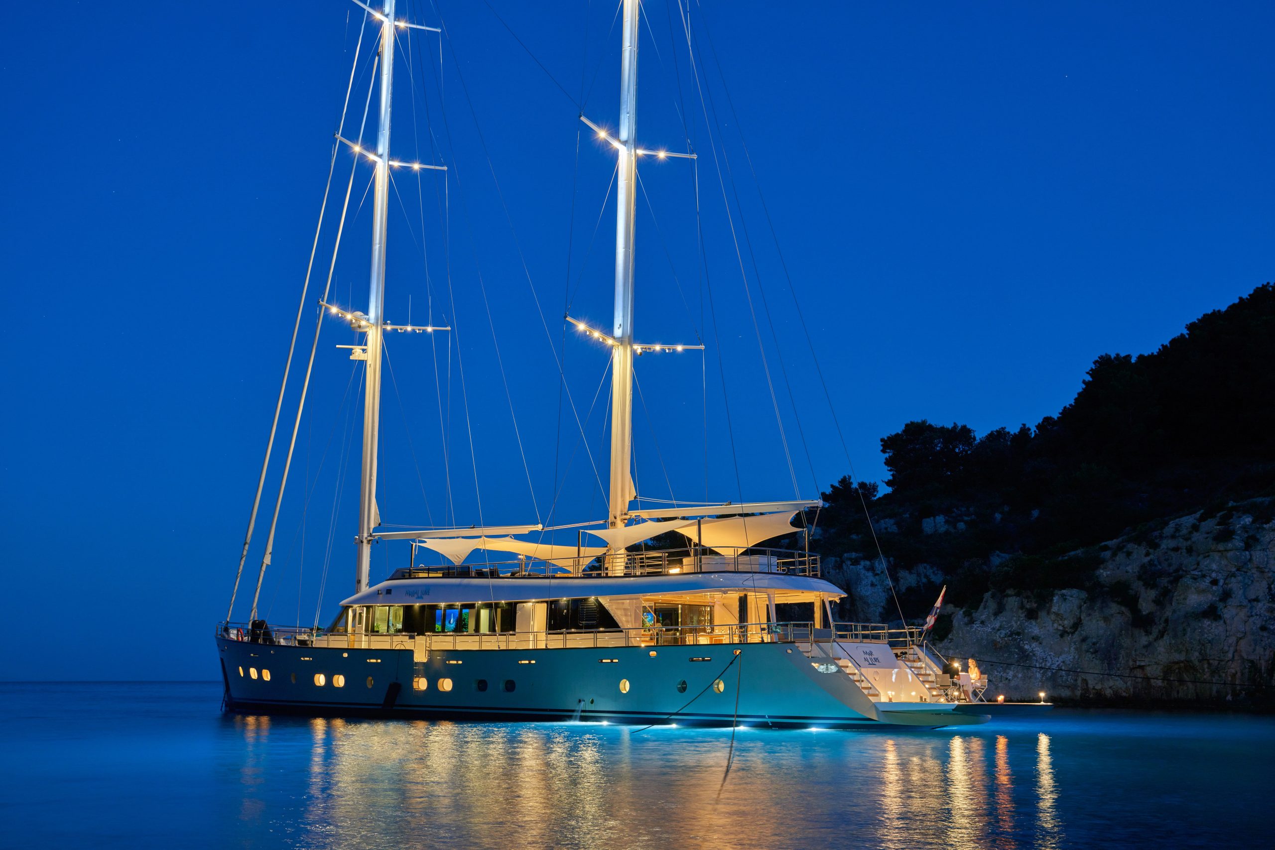 MARALLURE A large, modern yacht with tall masts is illuminated by lights at dusk, anchored near a rocky shoreline with trees, reflecting softly on calm blue water under a clear evening sky.