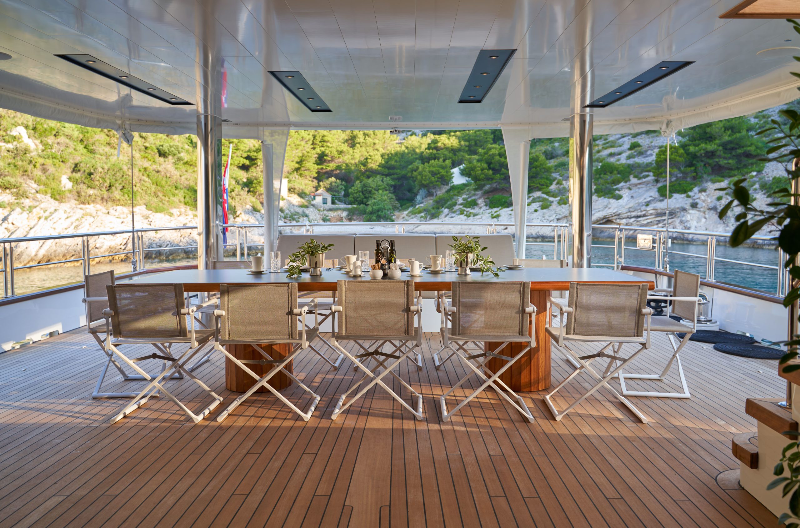 MARALLURE A large dining table with ten chairs is set on the deck of a yacht, overlooking calm water and a rocky, tree-covered shoreline. The table is set for a meal, with sunlight streaming onto the wooden deck.