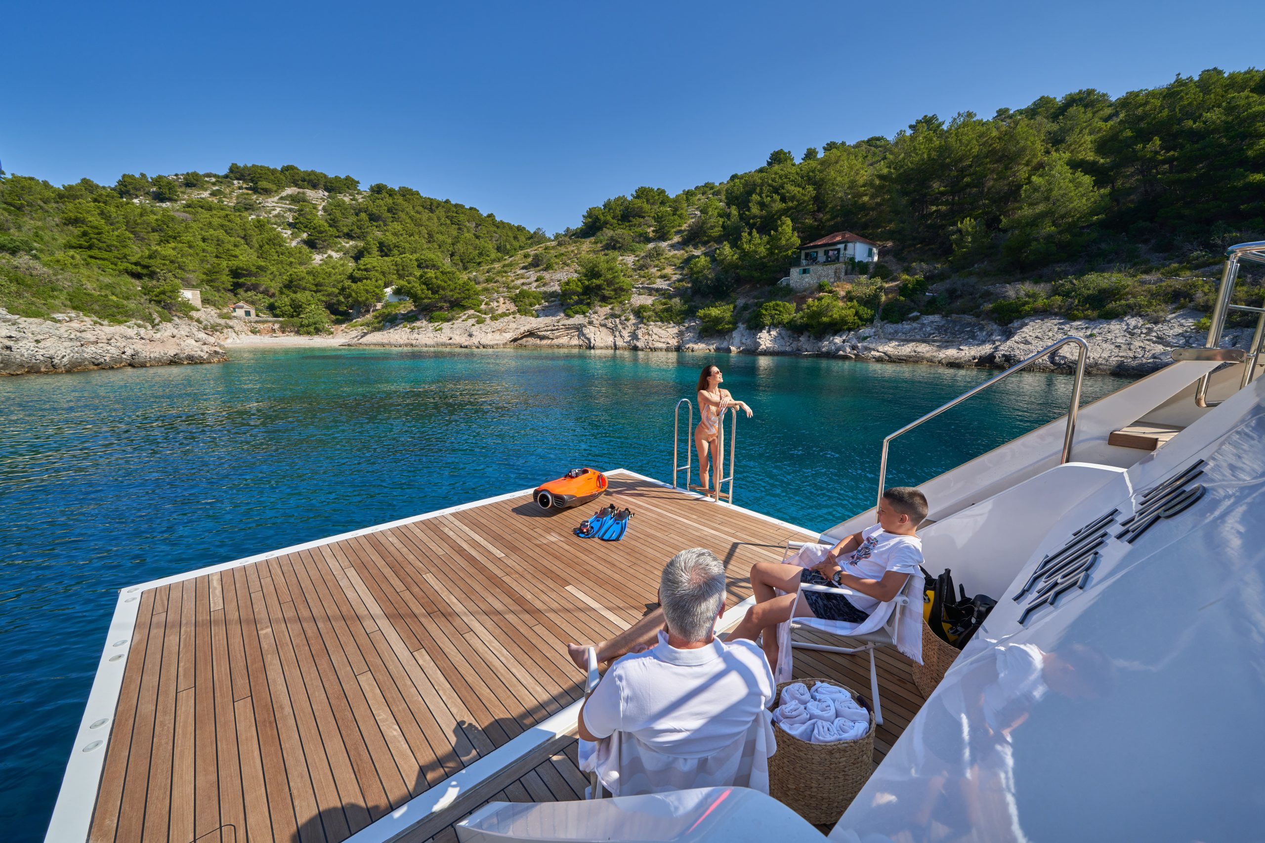 MARALLURE Two people relax on lounge chairs on a yacht deck, while one person stands near the waters edge. The yacht is anchored in a calm, clear blue bay surrounded by rocky hills and green trees under a clear sky.