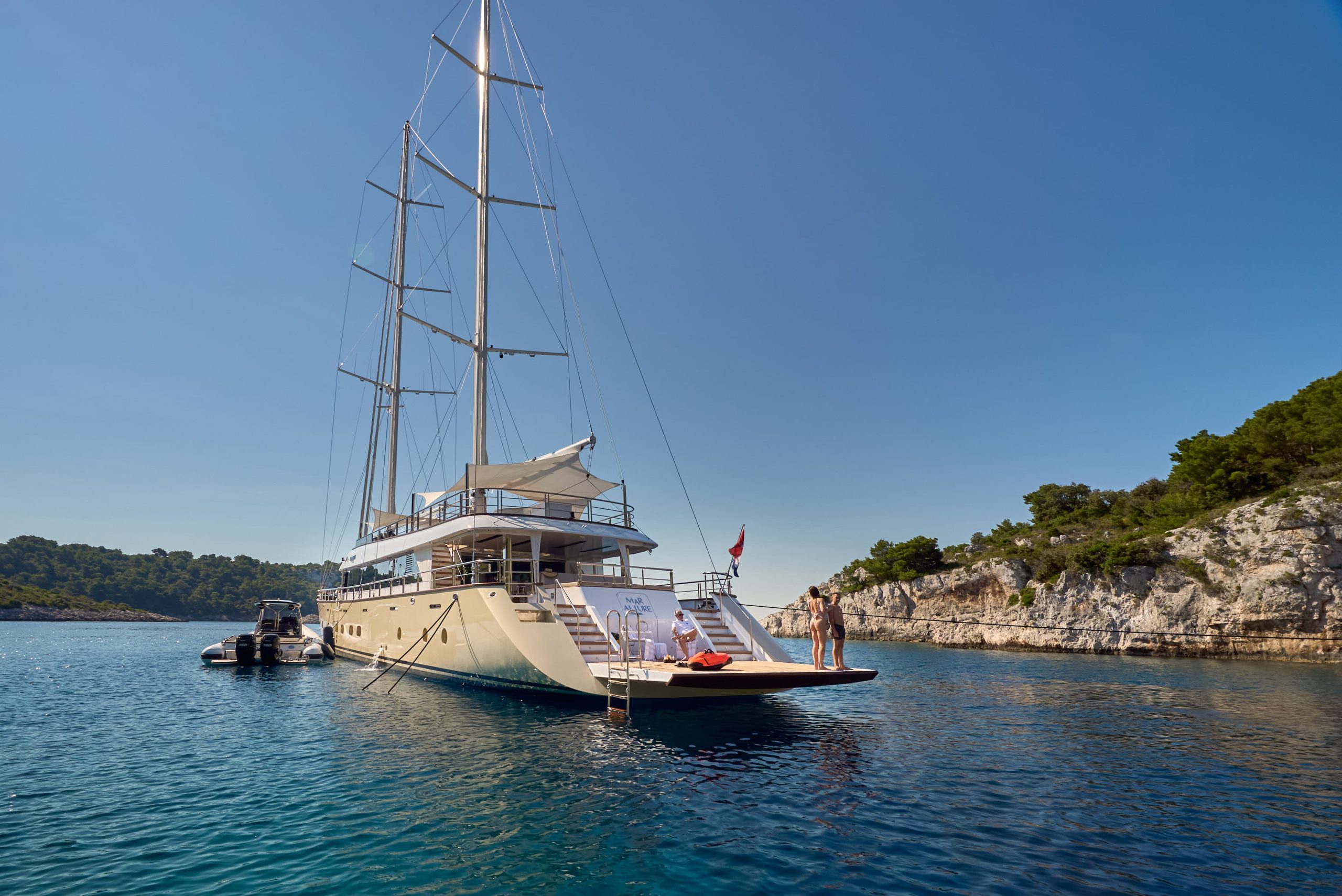 MARALLURE A large luxury yacht anchored in clear blue water near a rocky, tree-lined coastline. Two people stand on the yachts rear deck, and a small boat is tied alongside. The sky is clear and sunny.