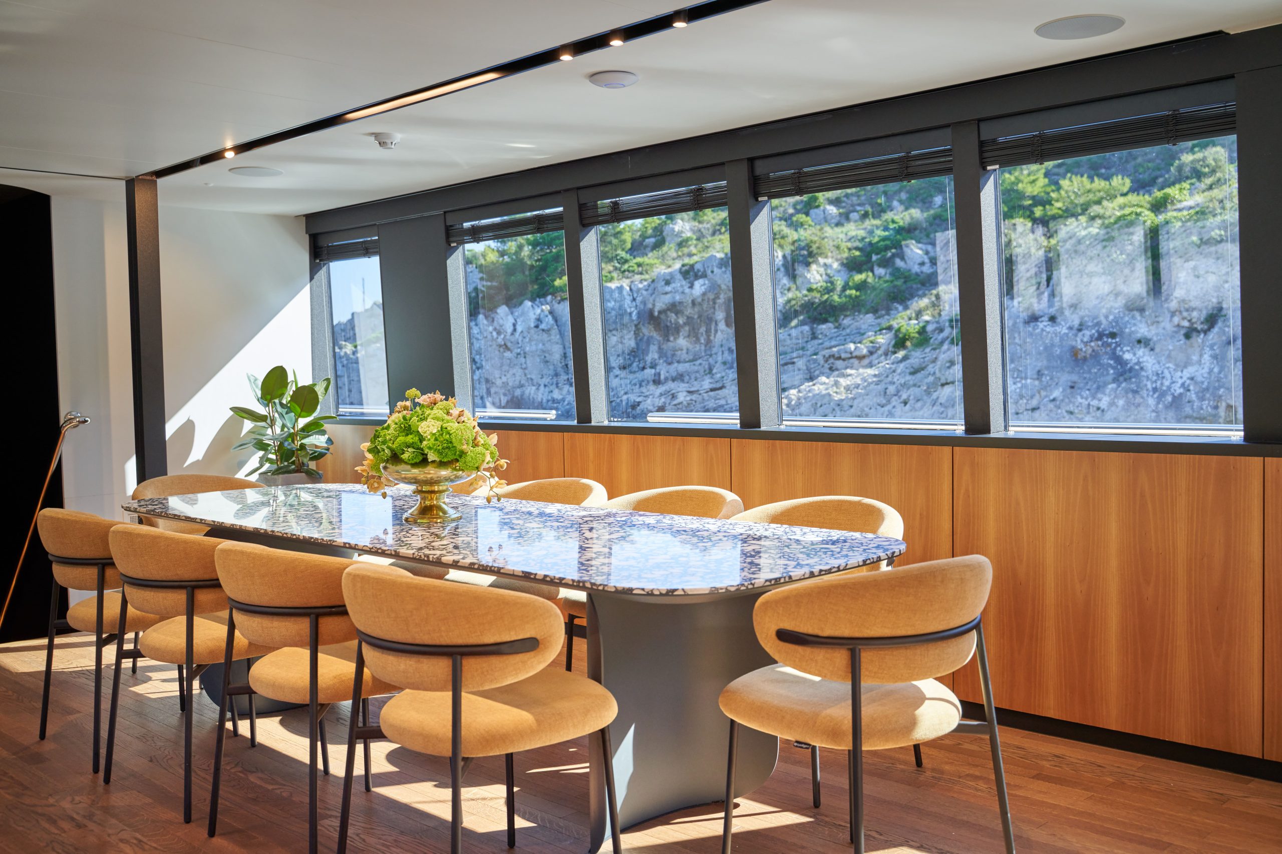 MARALLURE A modern dining room with a marble table, eight beige chairs, and large windows overlooking a rocky hillside. Sunlight brightens the space, which features wooden floors, a plant, and a flower centerpiece on the table.