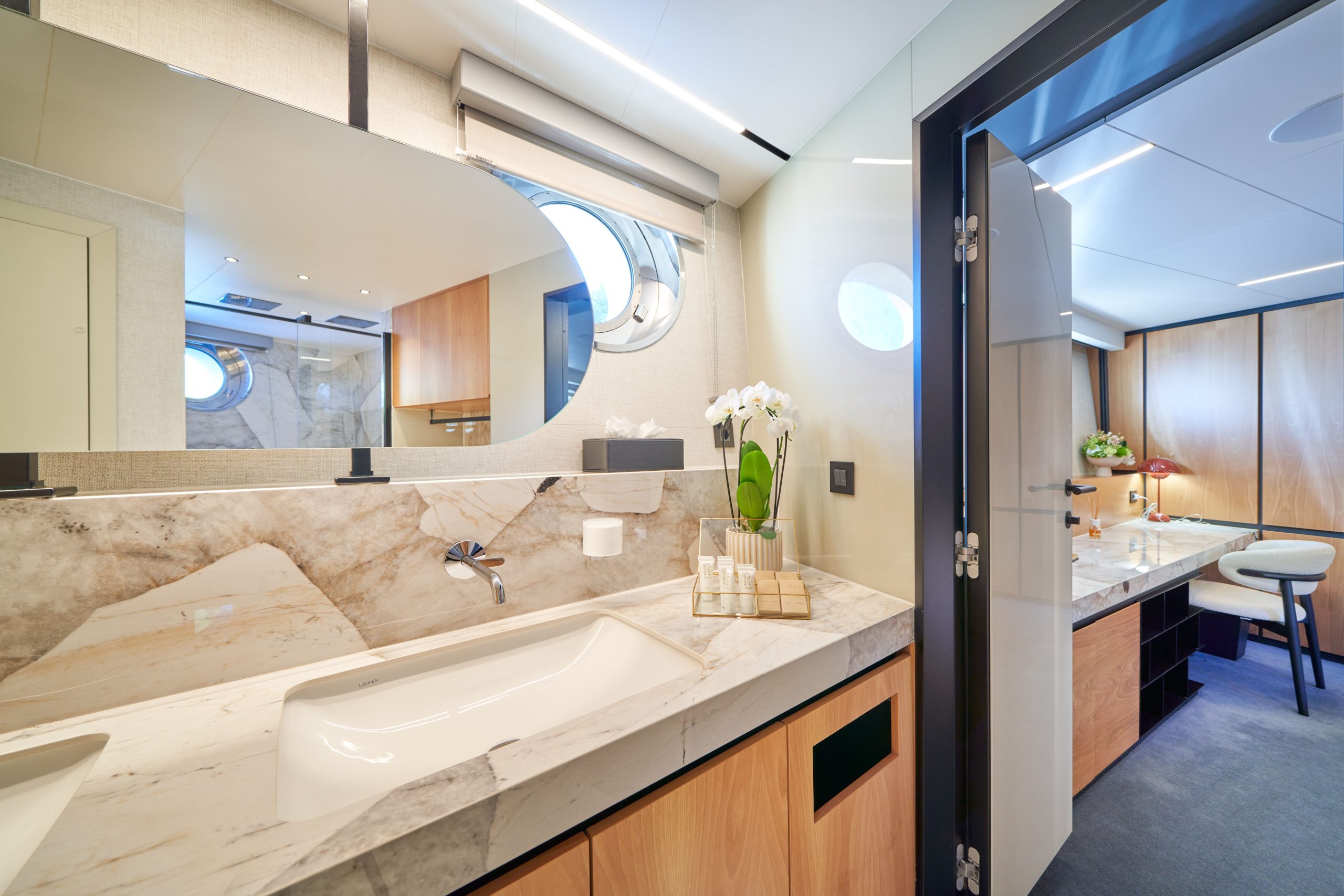 MARALLURE A modern bathroom with a marble countertop, white sink, large mirror, and porthole window. Visible through an open door is a study area with a desk, chair, and wood-panel walls. Orchids decorate the sink area.