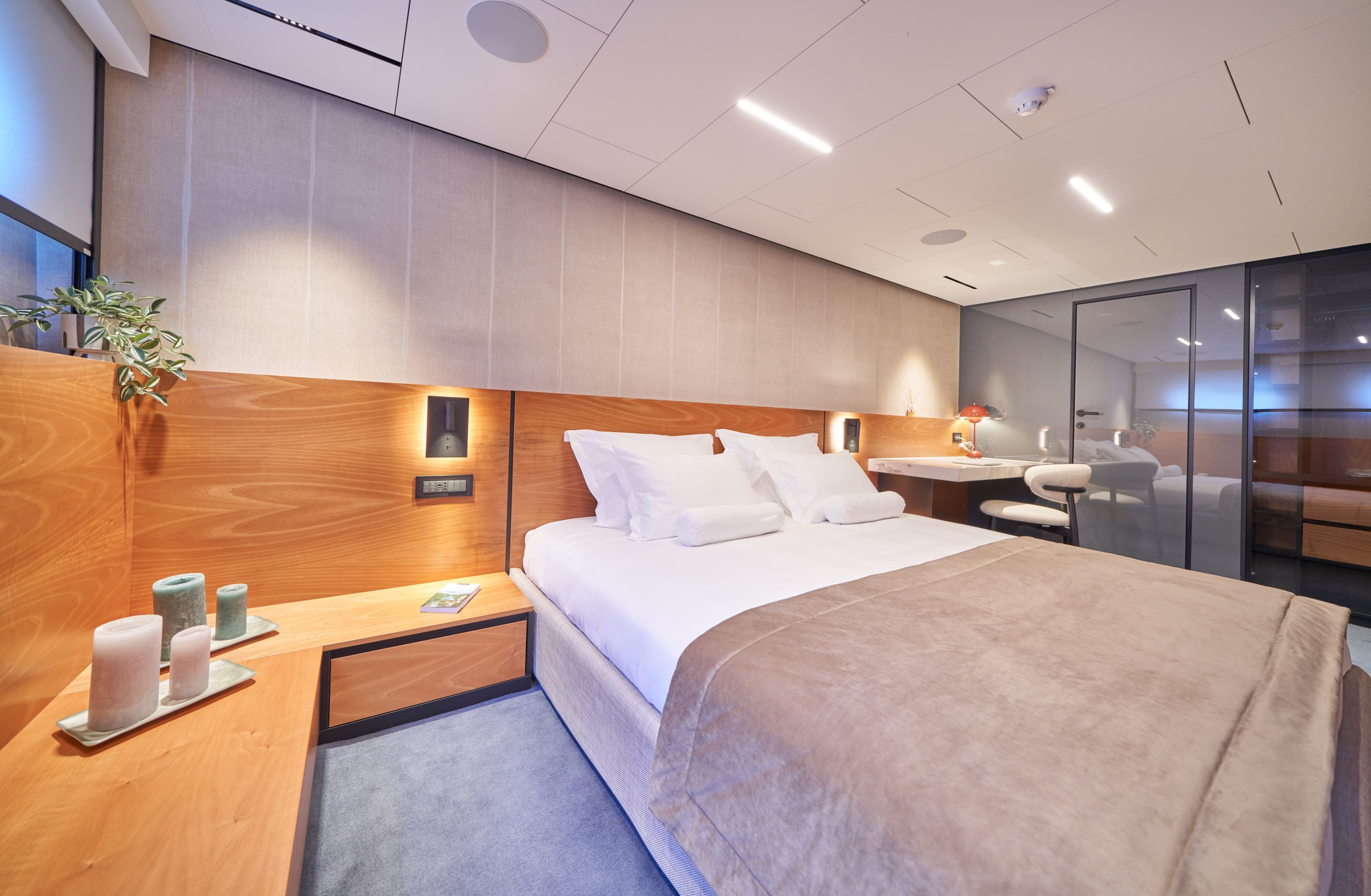 MARALLURE A modern hotel room with a neatly made double bed, beige linens, wooden headboard, bedside table with candles, and a desk with a chair and a lamp in the background. The room is well-lit and stylishly decorated.