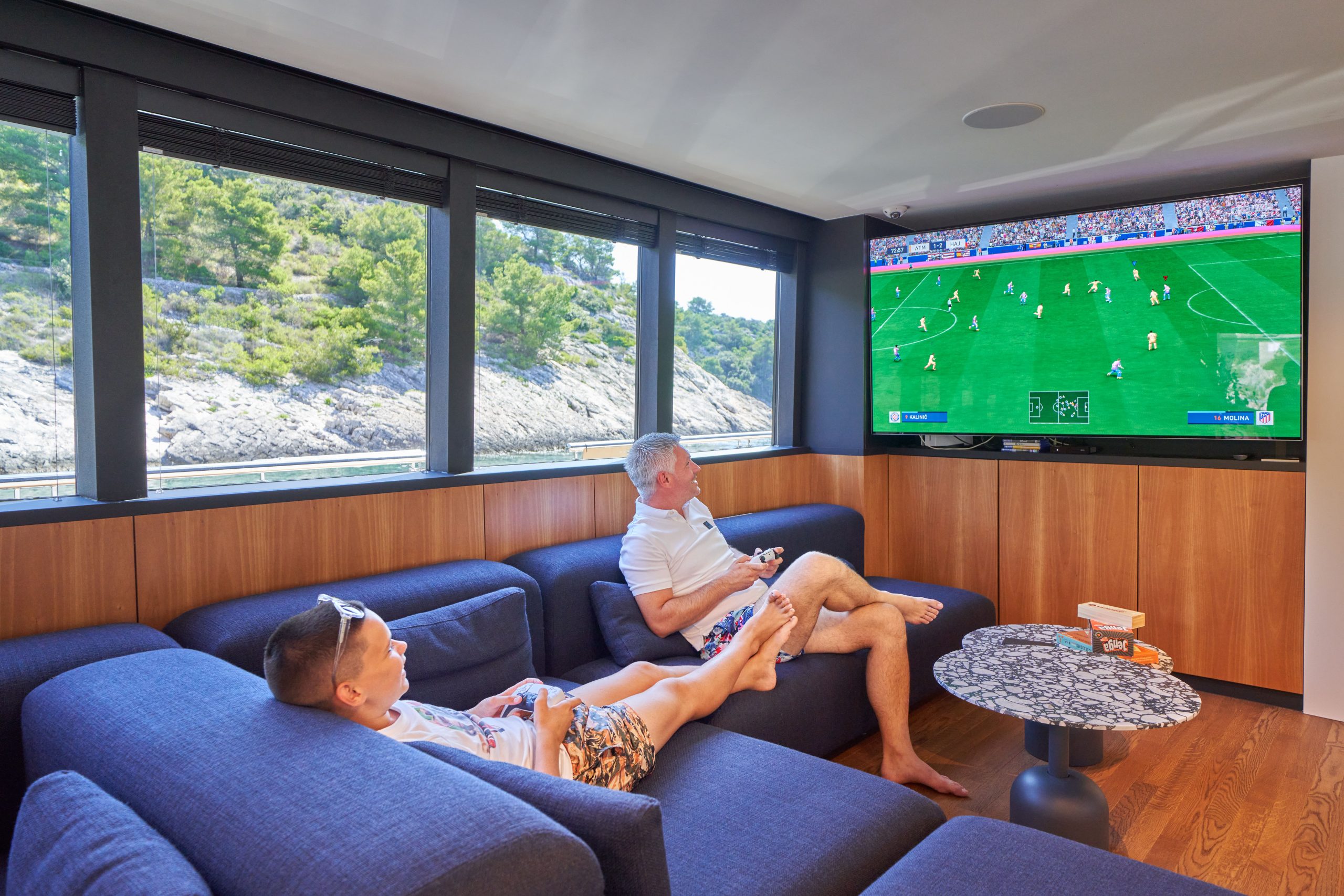 MARALLURE Two men relax on a blue sofa in a modern room with large windows, playing a soccer video game on a wall-mounted TV. Sunlight streams in, and a scenic rocky landscape is visible outside.