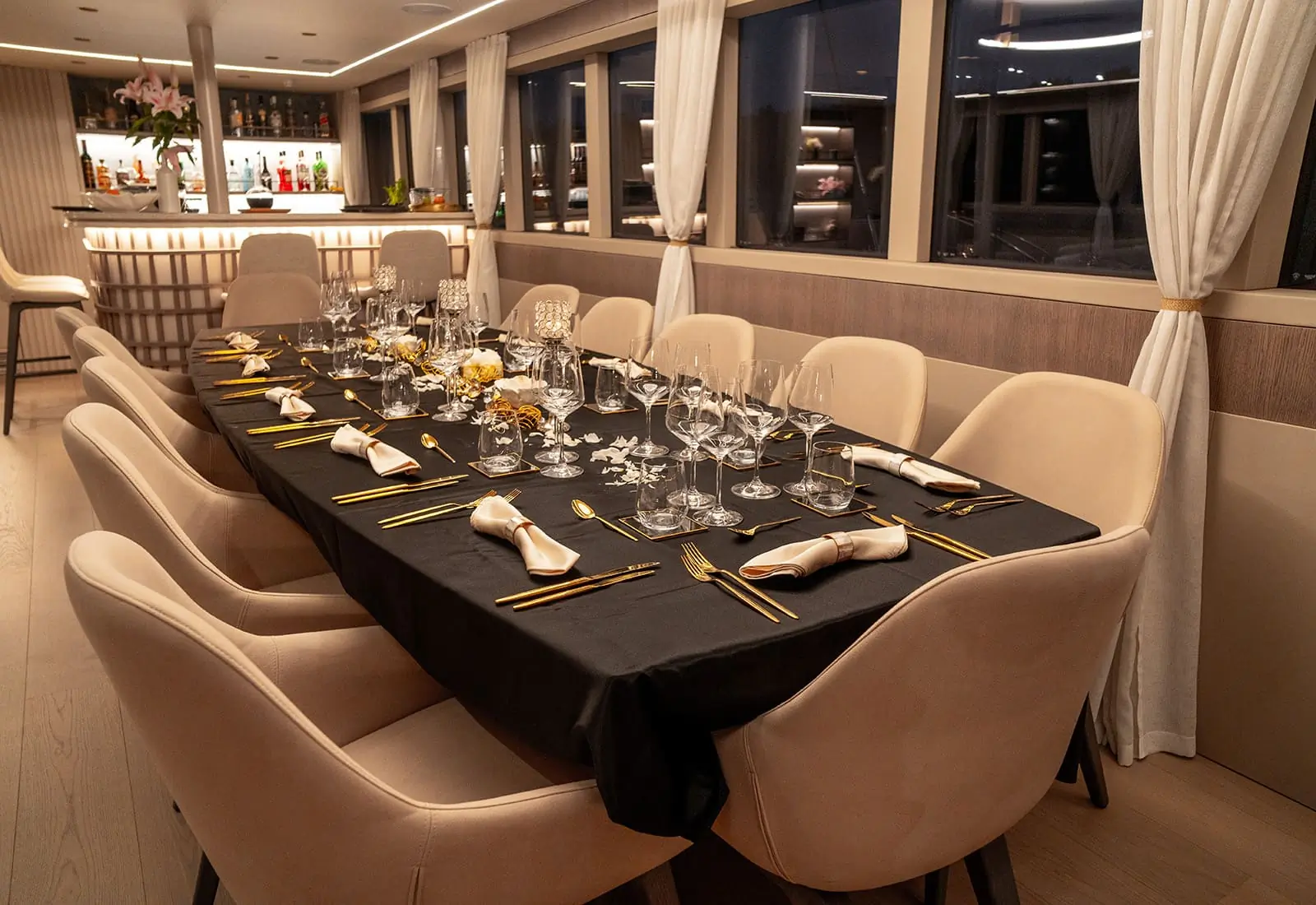 Elegant dining room with a long table set for eight, featuring neatly arranged glasses, plates, gold cutlery, napkins, and floral centerpieces. Neutral chairs surround the table; a bar is visible in the background.