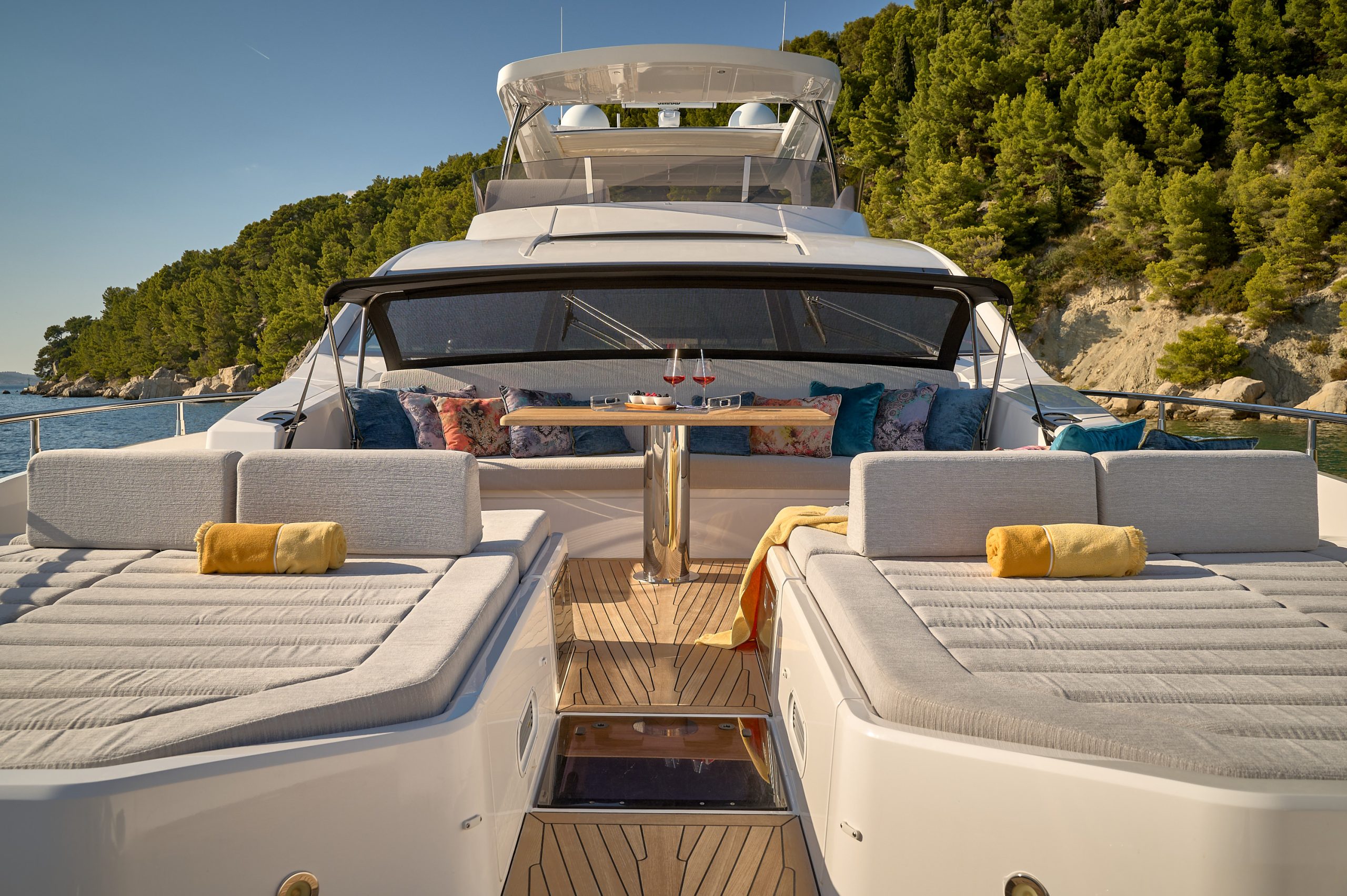 HIDEAWAY Luxurious yacht deck with cushioned lounge chairs, rolled yellow towels, a wooden table set with wine glasses, decorative pillows, and a scenic backdrop of trees and rocky shoreline under a clear sky.