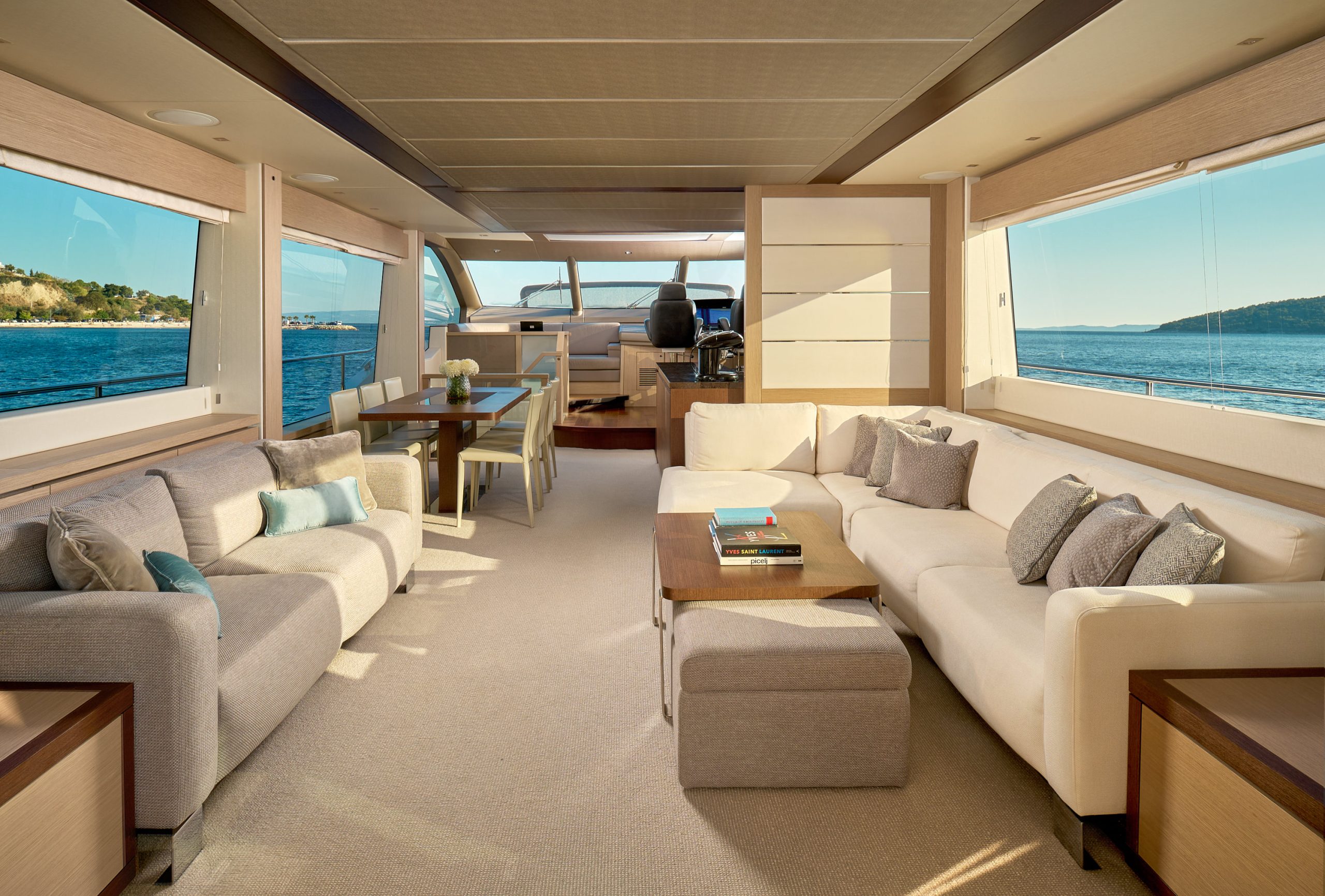 HIDEAWAY Spacious, modern yacht interior with large windows, beige and gray sofas, plush cushions, a coffee table with books, a dining area, and views of the blue ocean and distant hills through the windows.