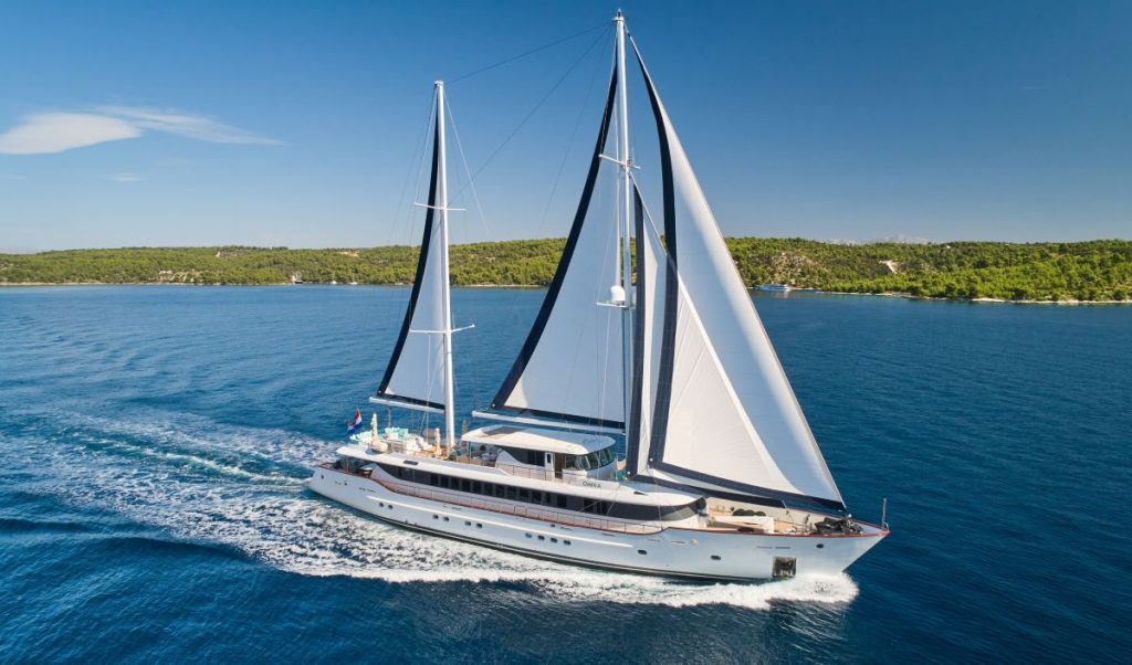 OMNIA A large white sailing yacht with two tall masts glides across calm blue water near a green, forested shoreline under a clear blue sky.