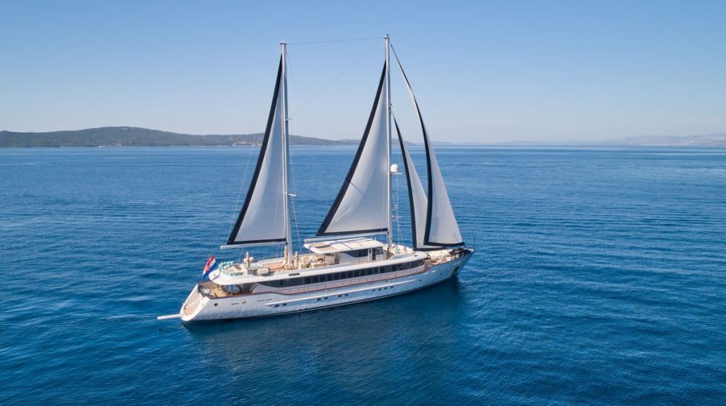 OMNIA A large, elegant sailing yacht with two tall masts and white sails glides on calm blue water under a clear sky, with distant green hills visible on the horizon.