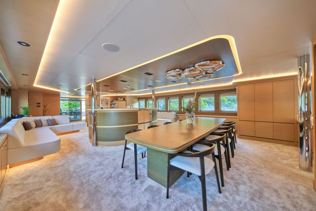 OMNIA A spacious, modern yacht interior featuring a dining table with six chairs, a plush carpet, built-in cabinetry, large windows with scenic views, and a curved white sofa. Warm lighting highlights the elegant design.