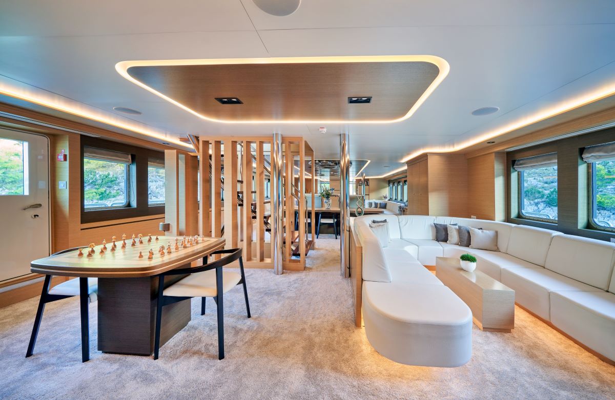 OMNIA Luxurious yacht interior with modern decor, light wood finishes, a large curved white sofa, a table set up for chess, soft beige carpeting, divided spaces, and large windows showing scenic views outside.
