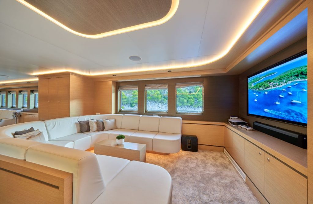 OMNIA A modern yacht lounge features a large white sectional sofa, beige carpet, wooden walls, wide windows with ocean views, and a flat-screen TV displaying an aerial view of boats and blue water.