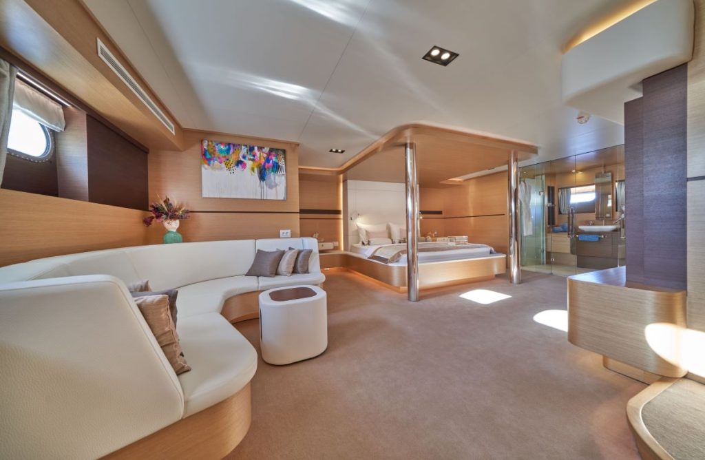 OMNIA Modern yacht interior with light wood finishes, a curved white sofa, small table, large bed, abstract artwork on the wall, and spacious layout with natural light from windows. A bathroom is visible in the background.