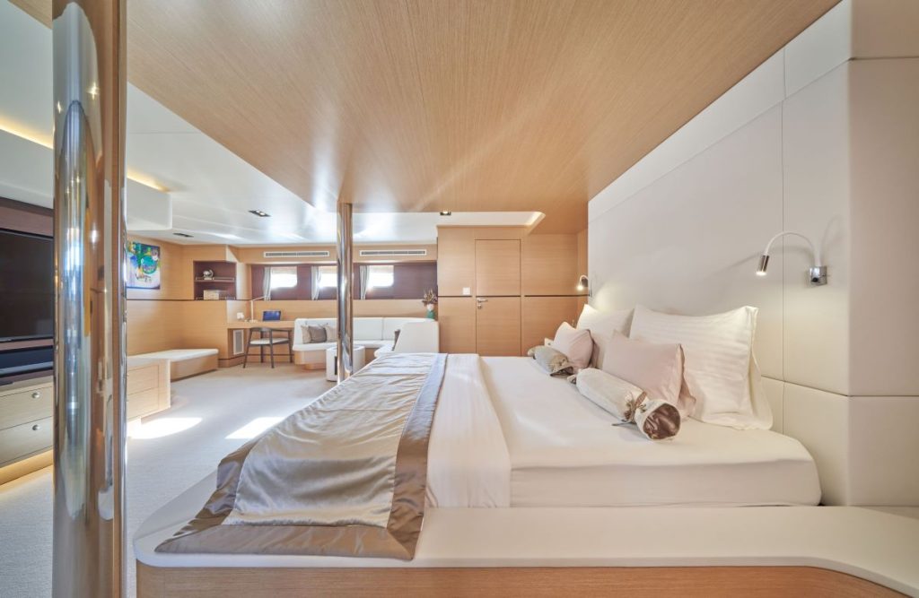 OMNIA A spacious, modern bedroom with light wood and white decor, featuring a large bed with white and beige linens, built-in lighting, a flat-screen TV, seating area, desk, and two small windows.