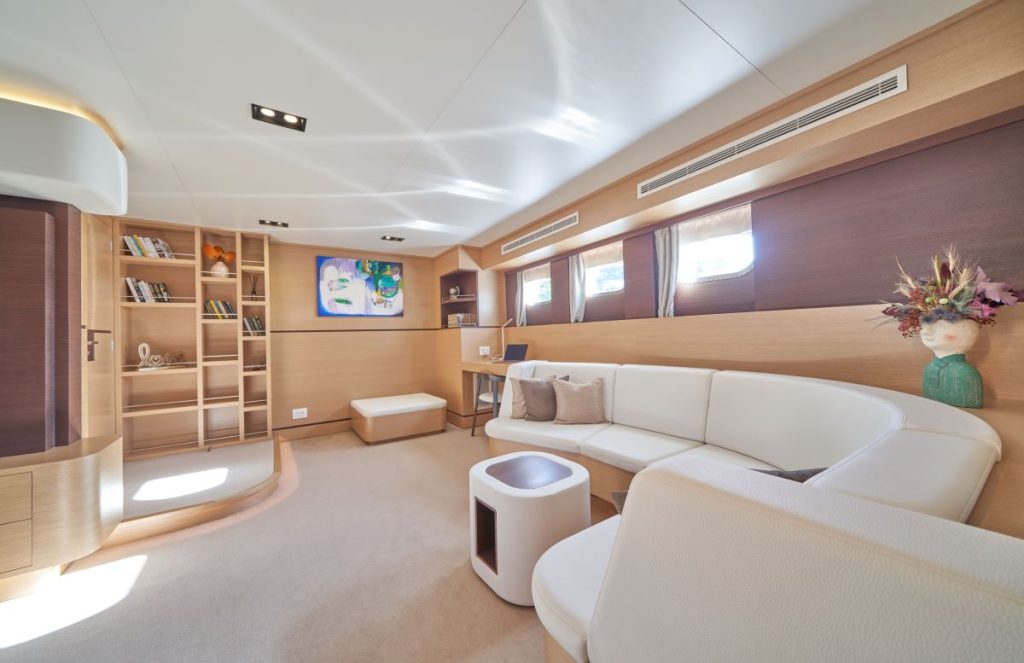 OMNIA Modern yacht interior with light wood paneling, white curved sofa, built-in bookshelves, and large windows letting in natural light. Decorative flowers and artwork add color to the minimalist, spacious lounge area.