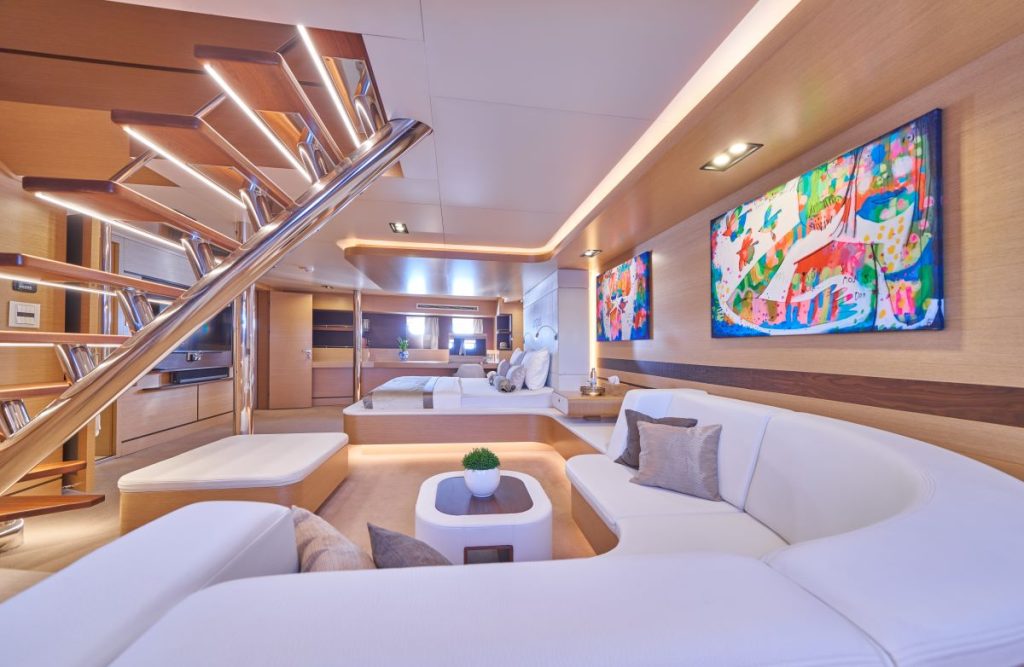 OMNIA Modern yacht interior with white curved sofa, colorful abstract artwork on the wall, a small table with a potted plant, wooden accents, and contemporary staircase with metal railings leading to an upper level.
