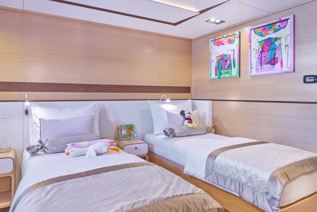 OMNIA A bright, modern bedroom with two single beds, each with beige and white bedding. Plush toys and children’s books are on the beds and nightstand. Colorful framed artwork hangs above the beds on the light wood-paneled wall.