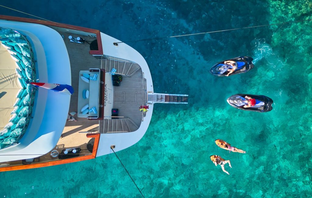 OMNIA Aerial view of a yacht anchored in clear blue water, with people swimming nearby and two jet skis floating beside them. The scene captures a sunny, tropical setting with vibrant, turquoise sea.