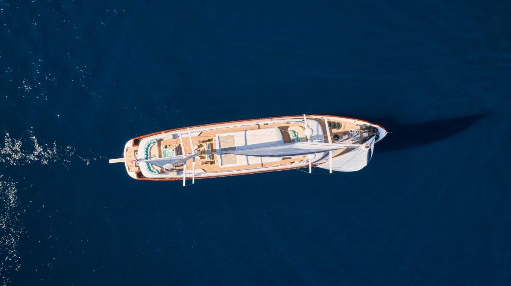 OMNIA Aerial view of a white sailboat with wooden decking cruising on deep blue water, creating a small wake behind it. The boat casts a shadow on the calm sea.