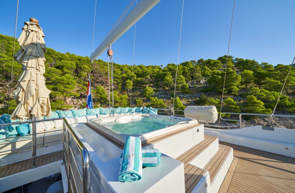 OMNIA A yacht deck featuring a hot tub with clear blue water, rolled-up towels, cushioned seating, and closed umbrellas, set against a backdrop of green trees and rocky shoreline under a clear blue sky.
