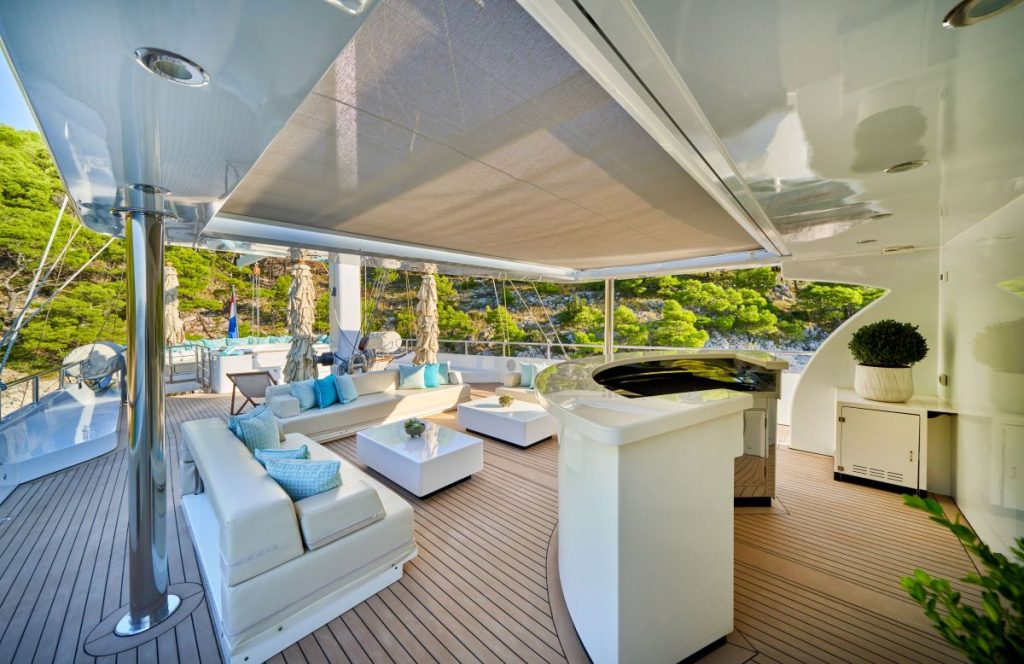 OMNIA Luxurious yacht deck with white sofas, tables, and a bar area under a canopy. Surrounded by lush green trees and calm water, the space is decorated with blue accents and plants, creating a relaxing outdoor lounge atmosphere.