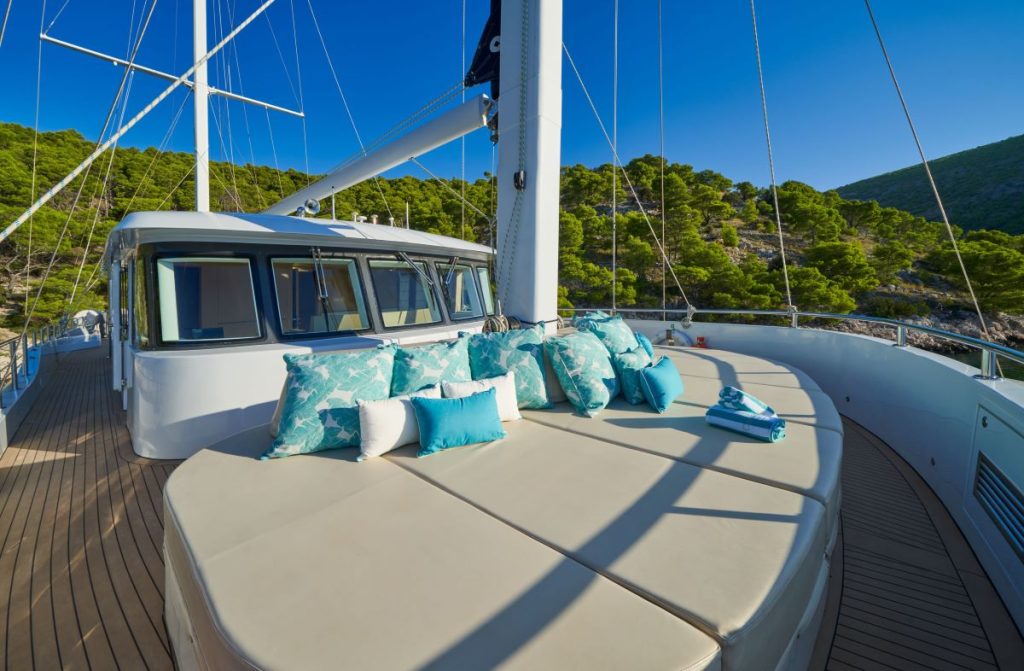 OMNIA Large sunbed on the deck of a yacht, decorated with blue and white patterned pillows and towels. Surrounded by trees and hills under a clear blue sky.