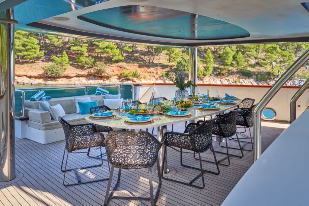 OMNIA A luxurious yacht deck features a set dining table for eight with blue and yellow tableware, wicker chairs, and a sofa seating area in the background; lush trees and water are visible beyond the railing.