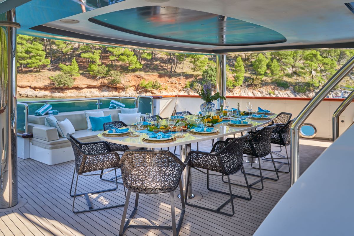 OMNIA A luxurious yacht deck features a set dining table for eight with blue and yellow tableware, wicker chairs, and a sofa seating area in the background; lush trees and water are visible beyond the railing.