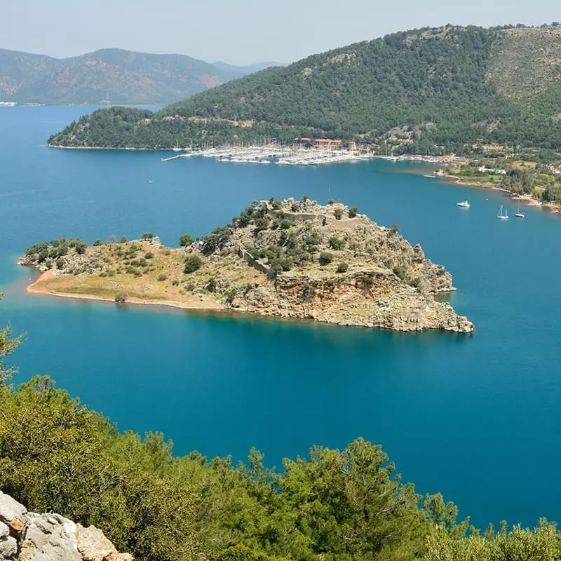 A rocky, green island sits in bright blue water, surrounded by hills covered with trees. Sailboats are docked nearby, and the sky is clear.