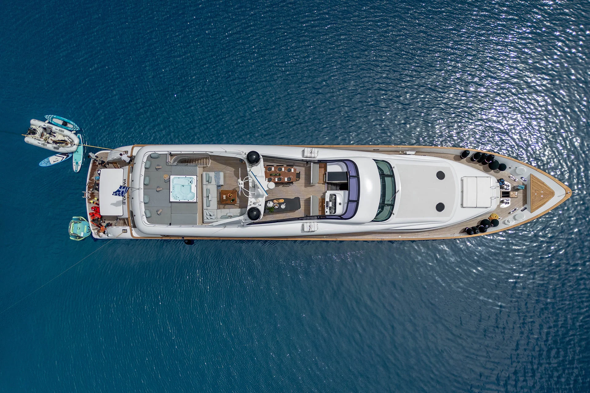 Aerial view of a large white yacht anchored in calm blue water, with smaller boats tied to its side and people visible on the deck.