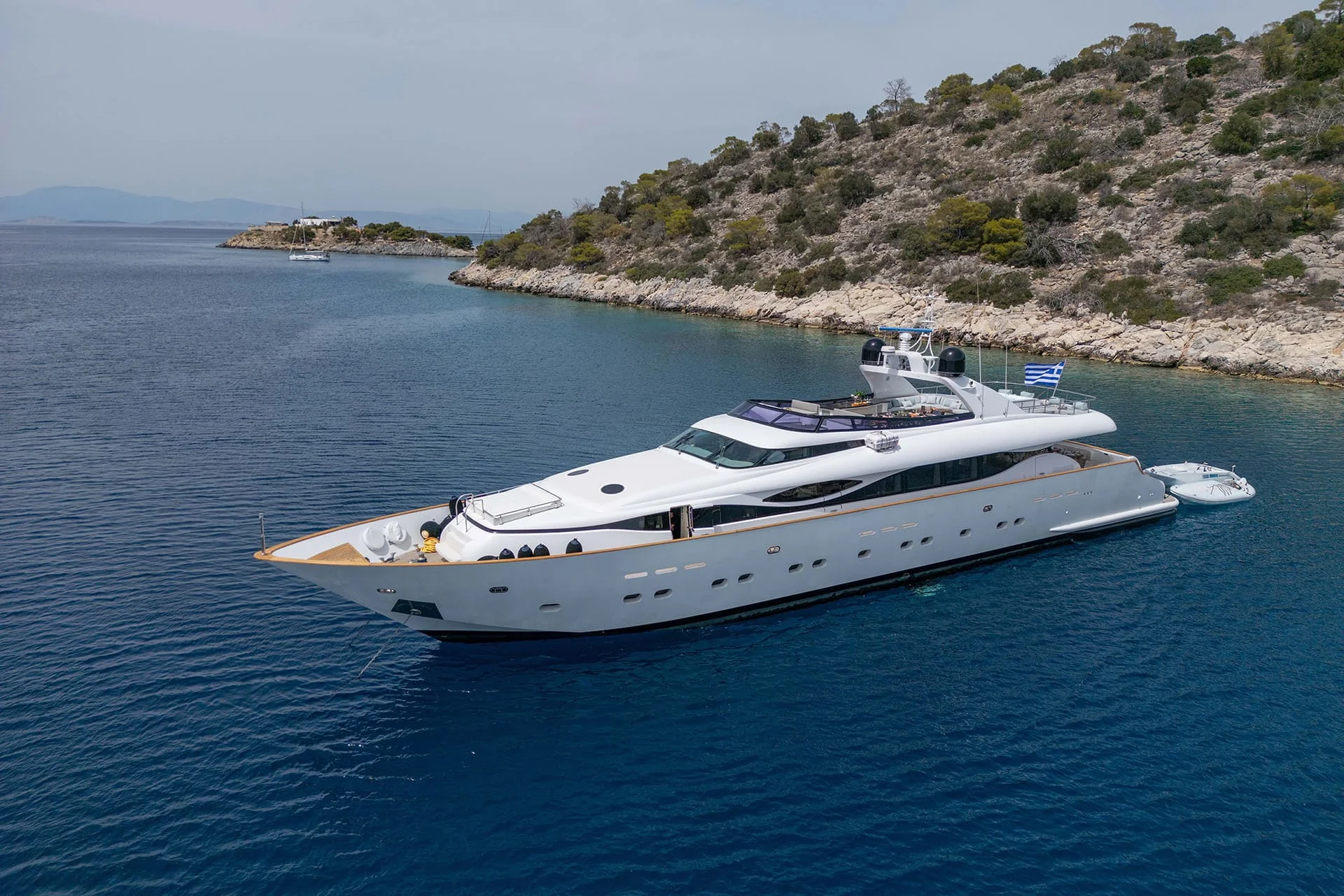 A large white luxury yacht floats on calm blue water near a rocky, tree-covered island under a clear sky. A small boat is tied to the yacht.