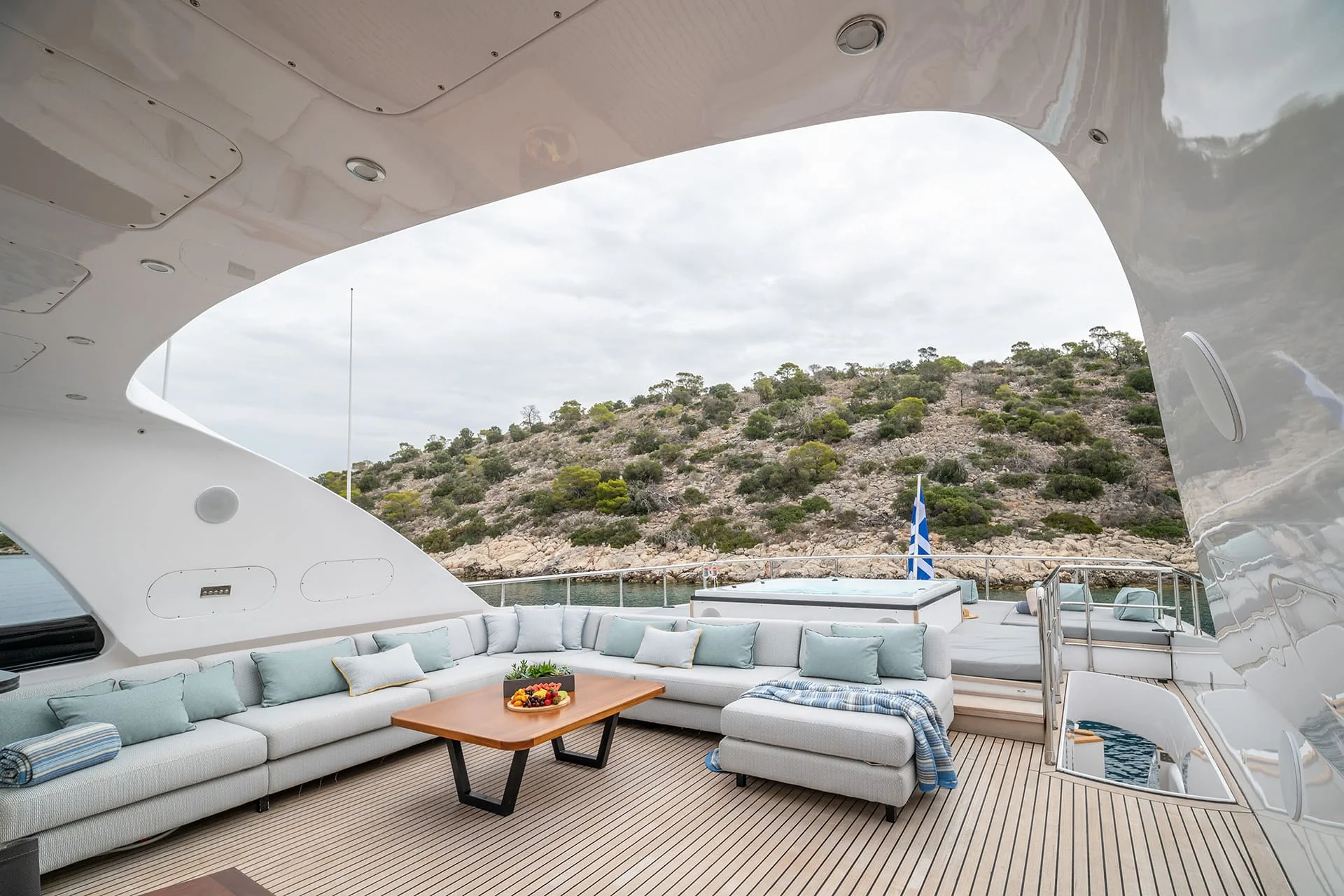 Luxurious yacht deck with a large cushioned seating area, wooden table with fruit, and a Greek flag in the background, overlooking a rocky, tree-covered hillside under a cloudy sky.