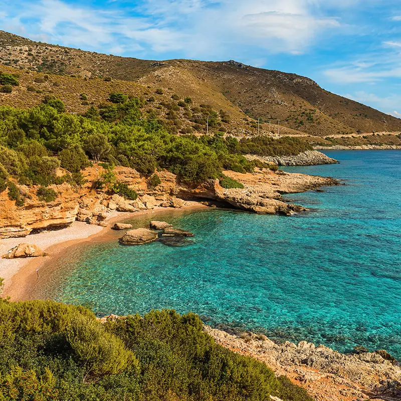 Rocky coastline with clear turquoise water, small sandy beach, and green shrubs. Hills rise in the background under a partly cloudy sky, creating a serene Mediterranean landscape.