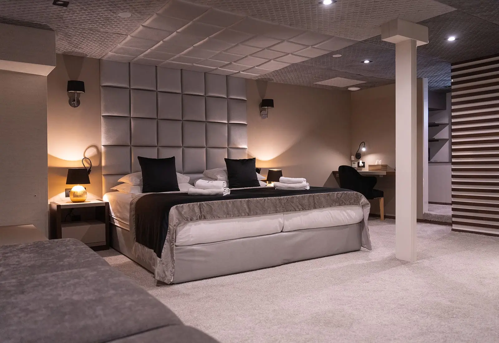 Modern hotel room with king-size bed, two black pillows, folded towels, bedside lamps, desk with chair, and soft gray carpet. The walls and ceiling have textured, padded panels for a cozy atmosphere.