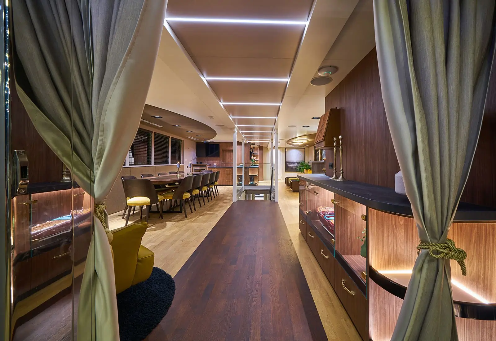 View of a luxurious, modern interior with wooden floors, elegant cabinetry, a dining area, and soft lighting, framed by curtains tied back with rope.