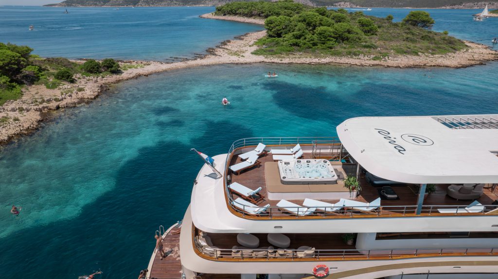A luxury yacht with a sun deck, lounge chairs, and a jacuzzi is anchored near a rocky island surrounded by clear blue water and lush greenery, with people swimming nearby.