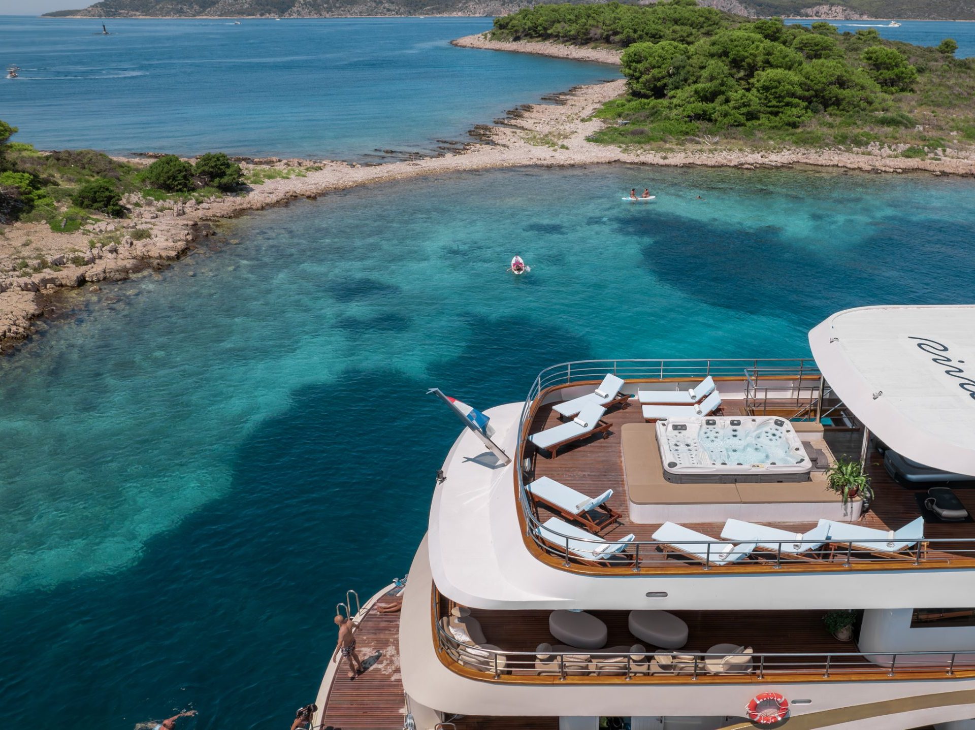 A luxury yacht with a sun deck, lounge chairs, and a jacuzzi is anchored near a rocky island surrounded by clear blue water and lush greenery, with people swimming nearby.
