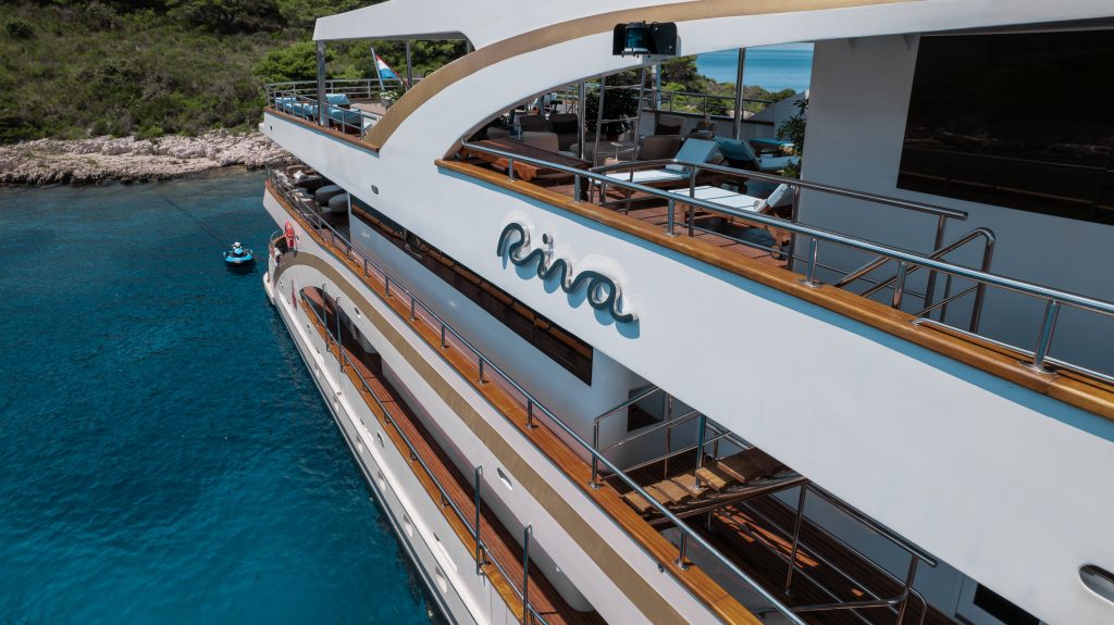 A close-up view of a luxury yacht named Riva docked in clear blue water near a rocky, tree-covered shoreline. The yacht has multiple decks with wooden railings and outdoor seating.