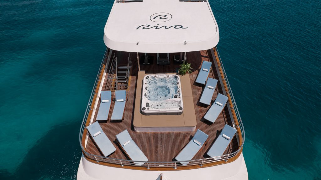 A luxury yacht deck with a central hot tub surrounded by cushioned lounge chairs overlooks clear blue ocean water. The word Riva is written on the white canopy at the top of the image.
