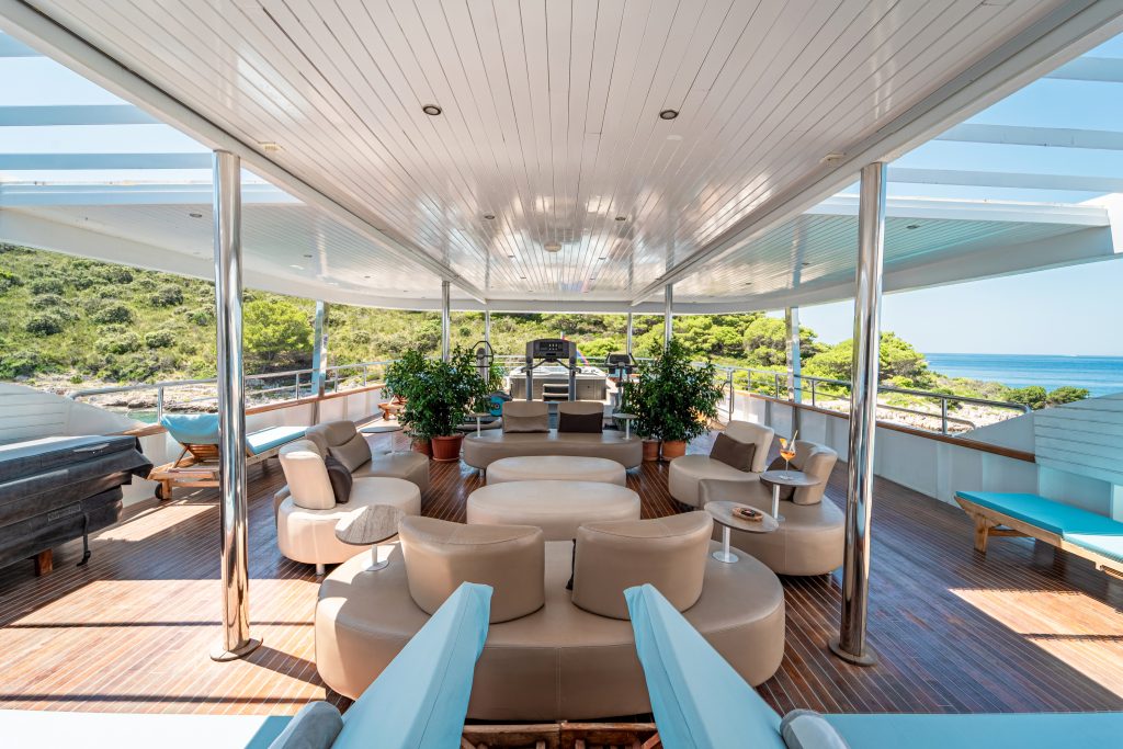 Spacious yacht deck with modern beige lounge chairs, small tables, potted plants, and wood flooring. Open sides reveal lush green trees and blue sea under a bright sky.