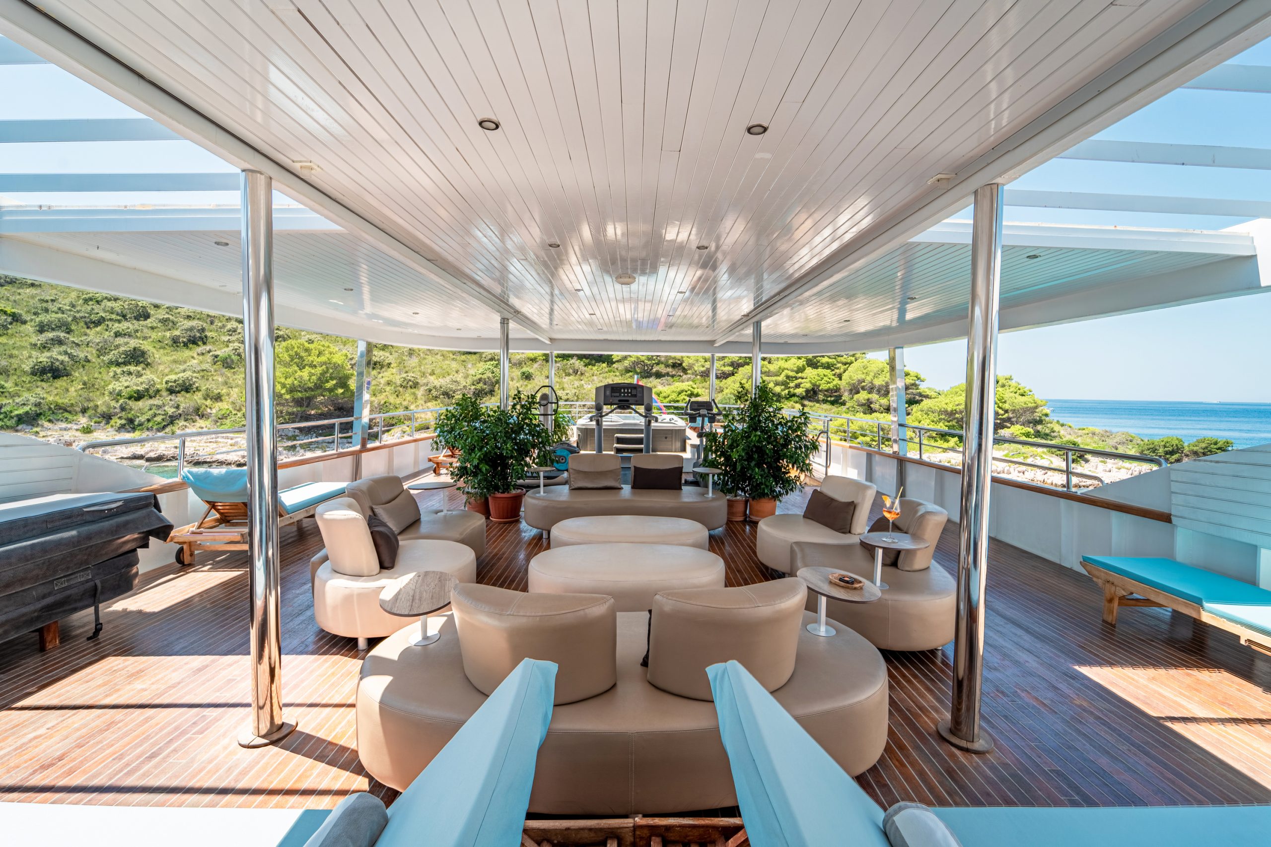 Spacious yacht deck with modern beige lounge chairs, small tables, potted plants, and wood flooring. Open sides reveal lush green trees and blue sea under a bright sky.