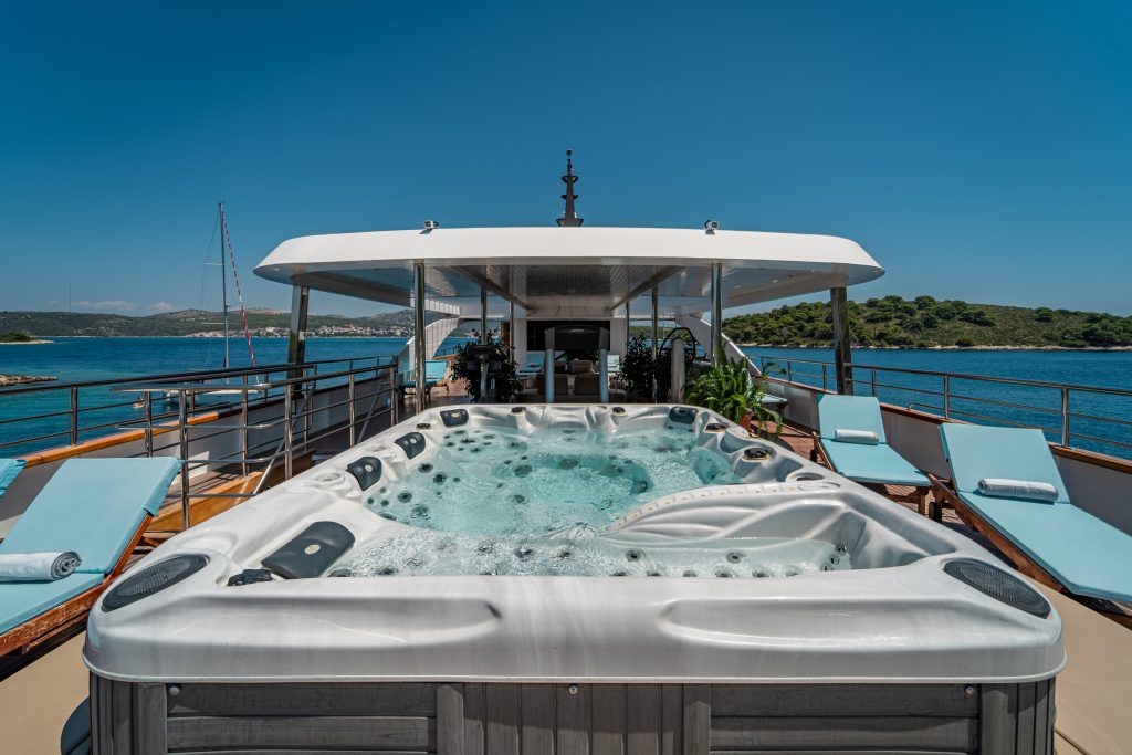 A luxurious yacht deck featuring a large bubbling hot tub, surrounded by sun loungers with blue cushions, overlooks clear blue water and a distant green shoreline under a clear sky.