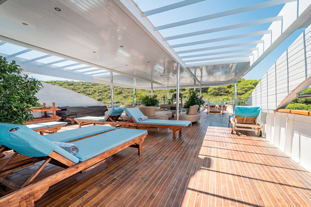 A spacious, covered outdoor lounge area with wooden flooring, blue cushioned sunbeds, lounge chairs, and potted plants, overlooking green hills under a clear sky.