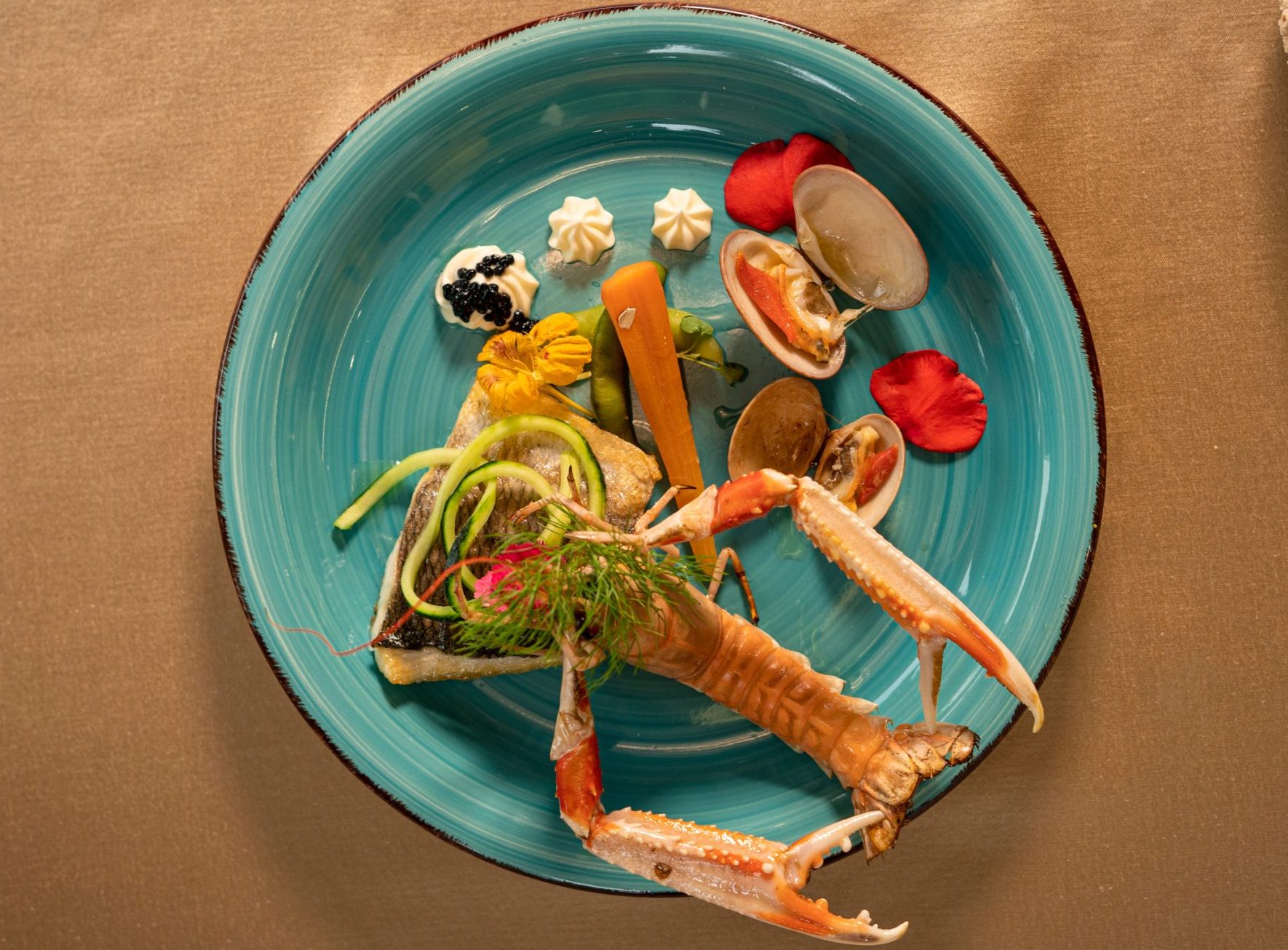 A turquoise plate with an artistic seafood dish, including crab claws, clams, mousse dollops, edible flowers, and vegetables, is arranged on a tan tablecloth with a decorative sea-inspired object to the side.