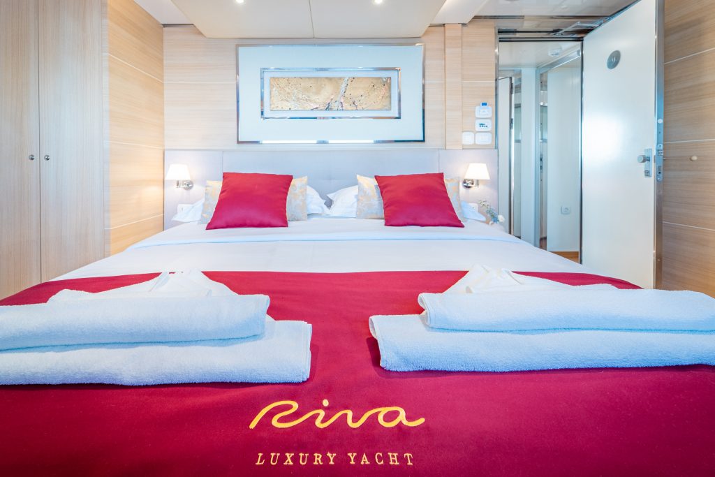 A neatly made bed with red pillows and a matching red blanket embroidered with Riva Luxury Yacht, two folded white towels at the foot, and light wood-paneled walls inside a modern yacht cabin.