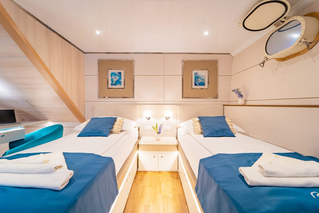 A bright, modern cabin with two single beds featuring blue bedding, a wooden floor, white walls, nautical decor, towels at the foot of each bed, and a round porthole window above one bed.